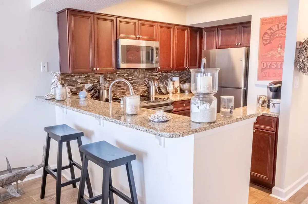 The kitchen features granite countertops, stainless steel appliances, wooden cabinetry, and glass jars for storage. The cozy setup suggests casual baking, hosting, and gathering, with a strong sense of warmth and togetherness.