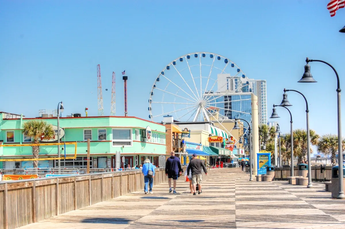 This vibrant exterior image showcases Myrtle Beach’s iconic ferris wheel, palm trees, colorful storefronts along the boardwalk, and people enjoying the lively atmosphere. The scene captures the playful, energetic spirit of the area and suggests a variety of fun activities like shopping, dining, and strolling by the ocean.