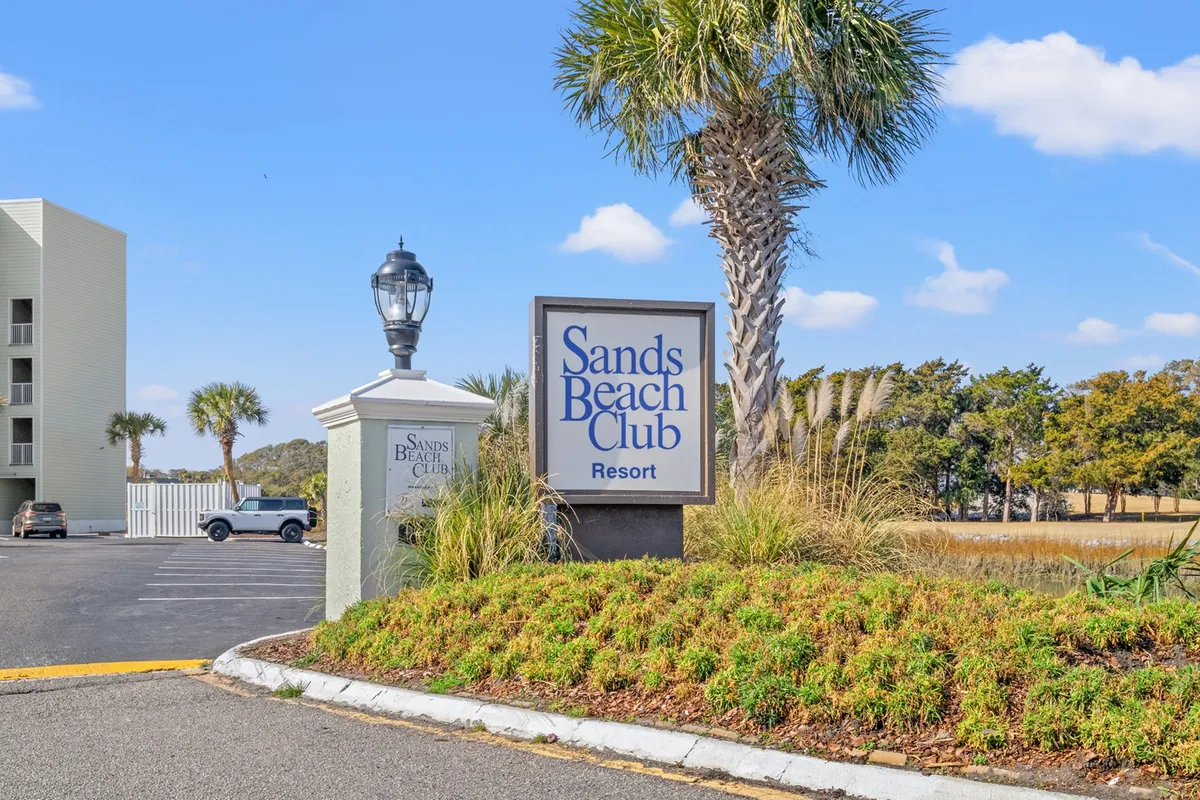 The Sands Beach Club sign is surrounded by lush palm trees and manicured landscaping, evoking a sense of arrival and resort relaxation. The setting suggests proximity to the beach and resort amenities in an inviting, relaxed atmosphere.