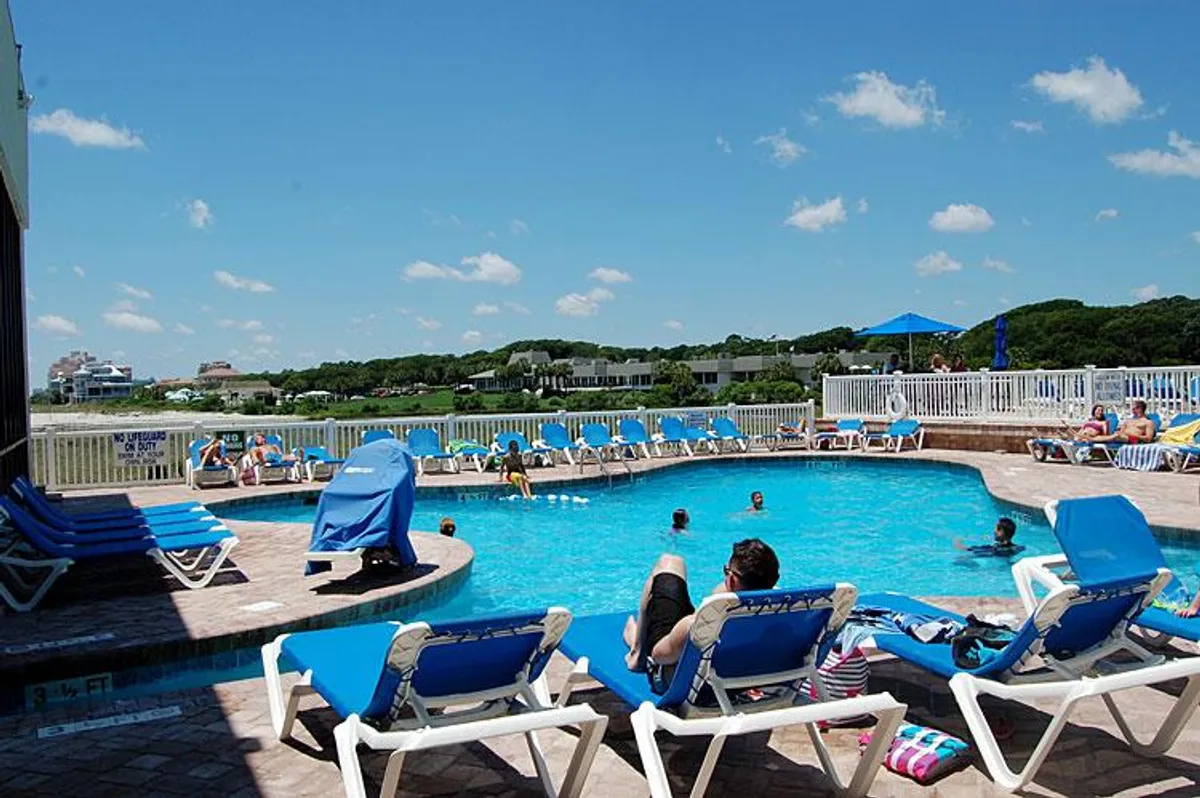 This vibrant exterior image captures the lively outdoor swimming pool area, complete with people swimming, lounging chairs, and umbrellas set against a backdrop of blue sky. The scene conveys a sense of relaxation, socialization, and the enjoyment of resort amenities.