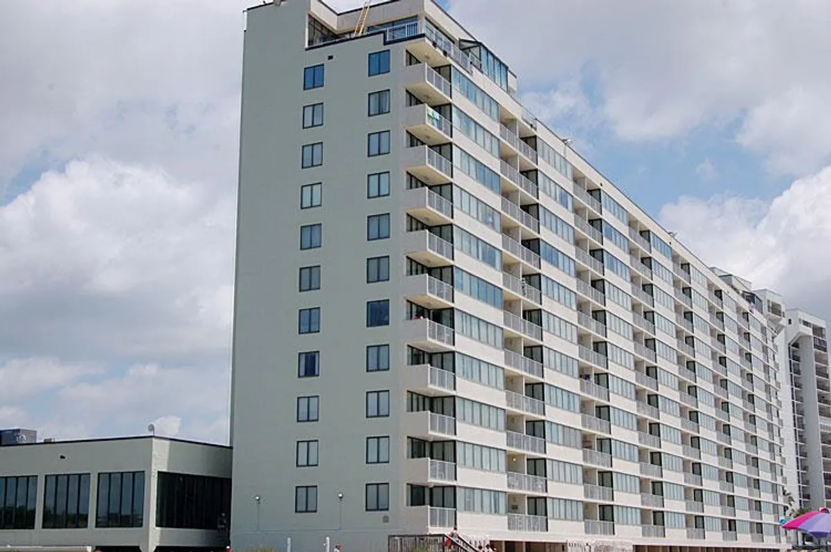 A modern exterior shot of Sands Beach Club reveals large windows, balconies, and people gathered on outdoor patios, capturing moments of relaxation, socializing, and enjoying views of Myrtle Beach. The building looks contemporary and welcoming, with hints of both excitement and comfort.