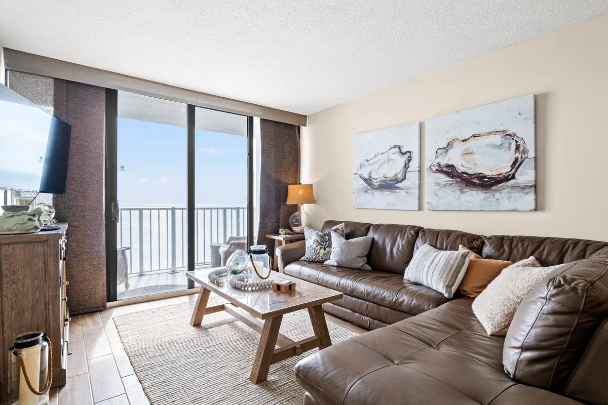 A cozy living room featuring a leather sofa, stylish coffee table, and artful paintings on the wall. Sunlight pours in through large glass doors, offering a direct view and access to the private balcony with the ocean beyond, seamlessly blending interior comfort with coastal scenery.
