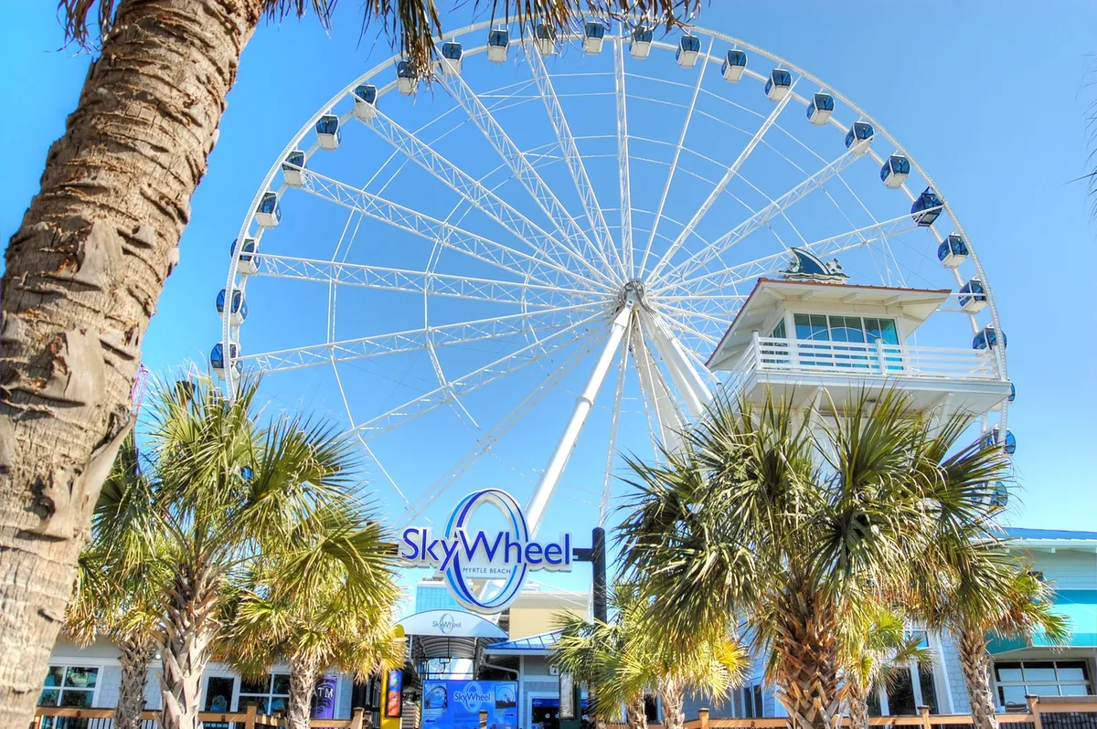 A dramatic shot of Myrtle Beach's iconic SkyWheel, surrounded by palm trees and with its entrance signage visible. The SkyWheel stands tall as a symbol of excitement and family-friendly attractions along the boardwalk.