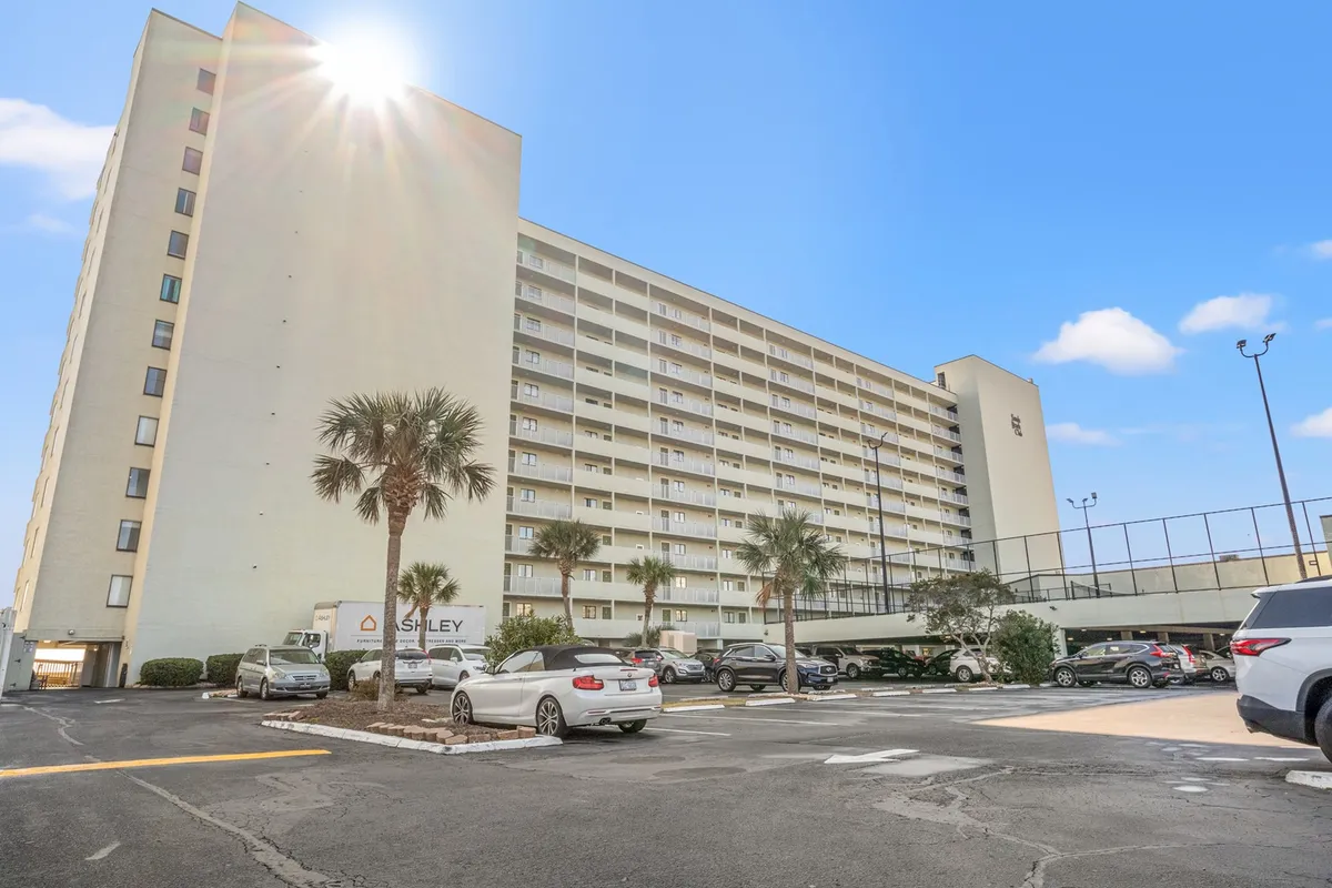 This image depicts the exterior of the Sands Beach Club building, framed by lush palm trees and a sunlit parking lot. The structure rises against a clear sky, evoking a sense of arrival and coastal relaxation. The palm trees lend a distinctly Southern atmosphere, reinforcing the Myrtle Beach location and the promise of mild winters.