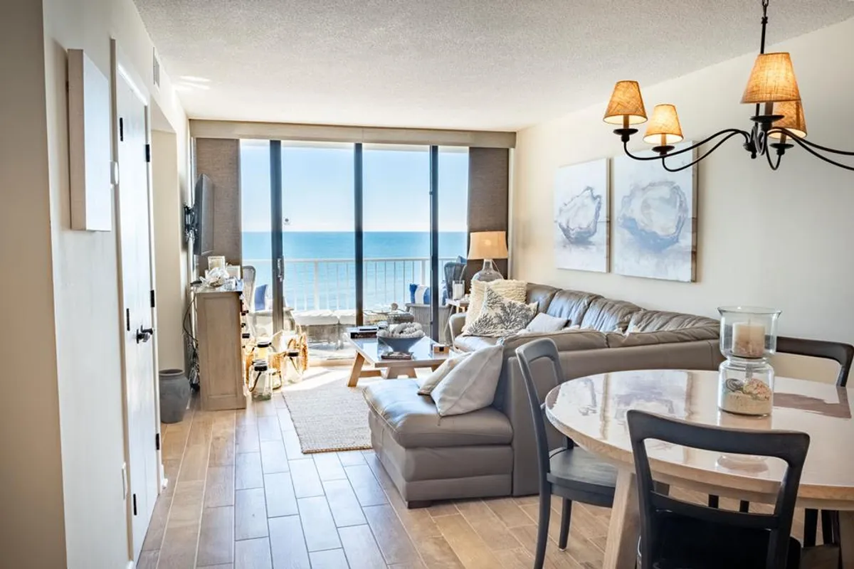 The image showcases a bright, spacious living room with a plush sofa and stylish dining table, all oriented toward expansive floor-to-ceiling windows framing a stunning panoramic ocean view. The scene exudes tranquility, with natural light flooding the space and the sparkling Atlantic just beyond the balcony, inviting guests to relax, dine, and soak up breathtaking sunrises without ever leaving the comfort of their condo.