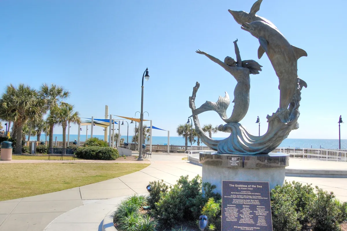This exterior shot showcases the iconic Goddess of the Sea sculpture overlooking the ocean, with dolphins and palm trees enhancing the lively waterfront scene. The broad promenade invites leisurely strolls, and the ocean view promises peaceful walks along the nearly empty winter beach.