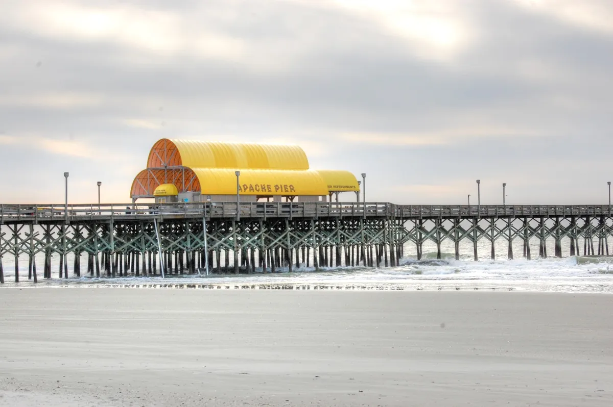 A scenic view of Apache Pier stretching into the ocean, with gentle waves below and a boardwalk inviting couples to stroll along the beach. The cloudy sky adds atmosphere, suggesting tranquility and the opportunity for peaceful seaside walks together.