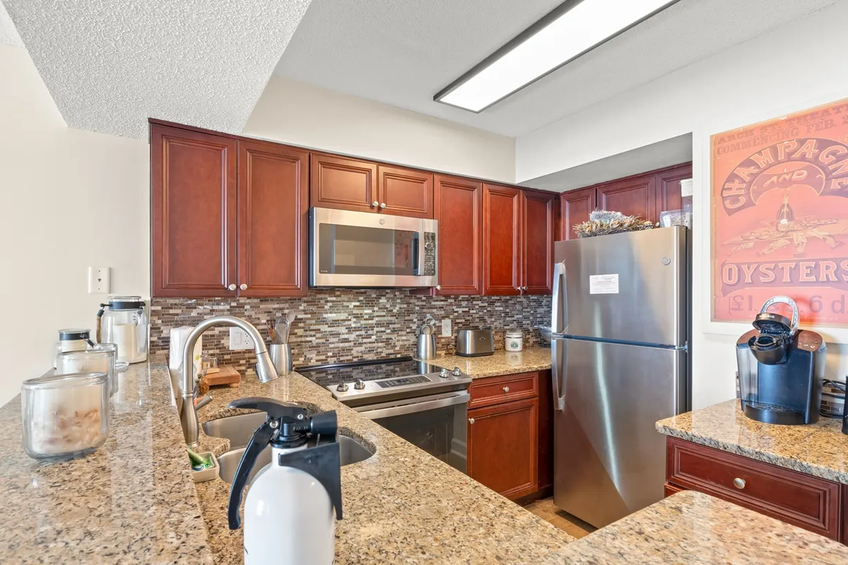 This image displays a spacious kitchen with granite countertops, stainless steel appliances including a microwave and dishwasher, wooden cabinetry, and decorative artwork. The kitchen setup conveys a welcoming atmosphere perfect for cooking, entertaining, or casual dining.